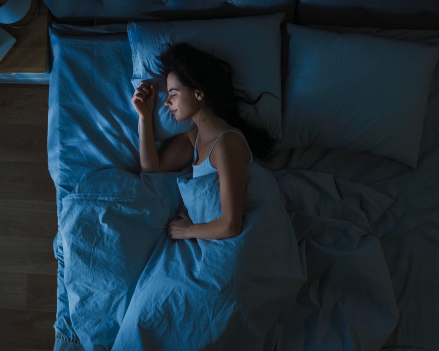 Top View of Beautiful Young Woman Sleeping Cozily on a Bed in His Bedroom at Night. Blue Nightly Colors with Cold Weak Lamppost Light Shining Through the Window.